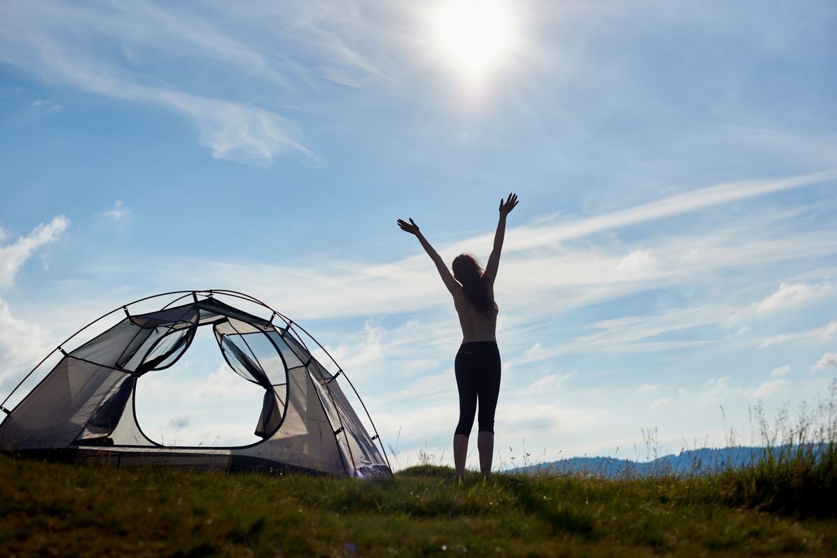 Back view of silhouette of sporty female tourist standing near the tent with lifting hands up in the air, enjoying summer morning in mountains. Camping lifestyle concept adventure vacations outdoor